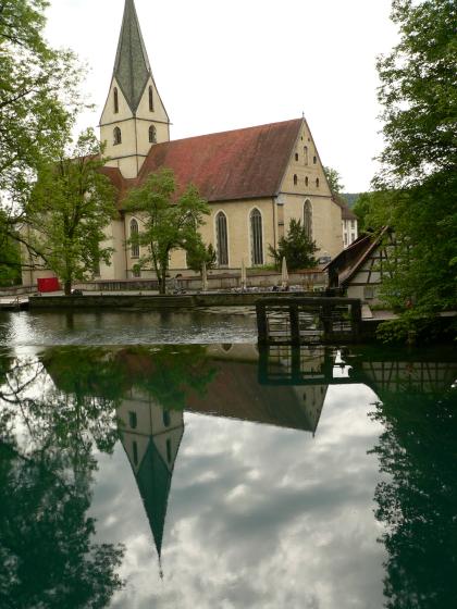 Eine schlichte, mit einem spitzen Turm versehene Kirche spiegelt sich in einer unmittelbar angrenzenden Wasserfläche.