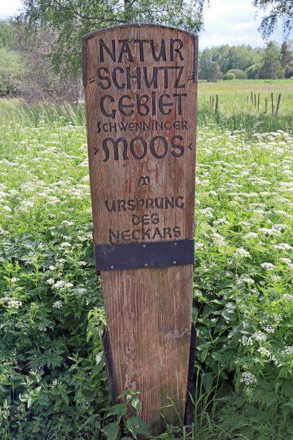 Blick auf eine schmale hohe Holztafel. Sie steht auf einer Wiese mit hohem Pflanzenwuchs und verweist auf das Naturschutzgebiet Schwenninger Moos sowie den Ursprung des Neckars.