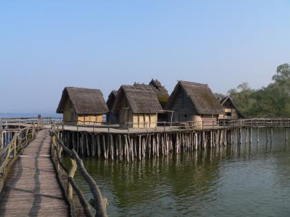 Das Foto zeigt mehrere Holz- und Lehmhütten, die auf einer von Holzpfählen gestützten Plattform stehen. Die Pfähle selbst sind im Grund eines großen Sees verankert. Ein Steg aus Holz führt zu den Hütten.