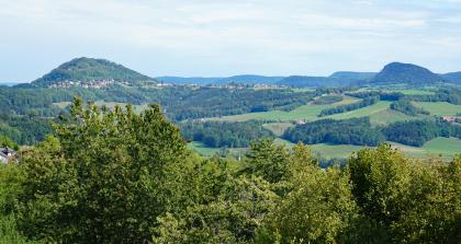Blick von erhöhtem Standort über Baumipfel auf eine hügelige, teils bewaldete Landschaft. Im Hintergrund links erhebt sich ein bewaldeter, von einer Ortschaft umgebener Bergkegel. Rechts außen sind zwei weitere, abgestufte Berge zu erkennen.