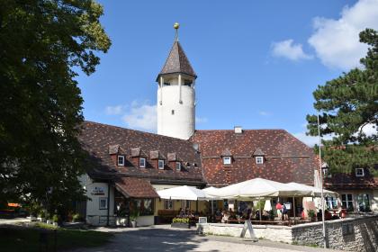Das Bild zeigt ein über Eck stehendes Gebäude mit kleinen Dachgauben. Im Eckpunkt steht ein runder weißer Turm mit spitzem Ziegeldach. Rechts vorne, auf einem Vorplatz, sind Sitzbänke, Tische und Sonnenschirme aufgestellt.