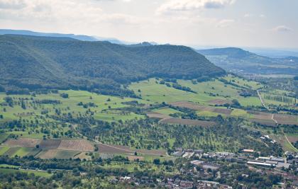 Blick von einem Aussichtspunkt auf eine nach links hin ansteigende Landschaft mit Wiesen, Äckern und einer Siedlung im flacheren Teil sowie bewaldeten Bergen, die auch im Hintergrund zu sehen sind.