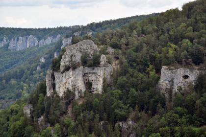 Blick auf mehrere große Felsformationen, die aus einem nach rechts ansteigenden, bewaldeten Berghang herausragen. Der größte Felsen, links der Bildmitte, hat eine kreisförmige Gestalt mit schroffen Wänden und oben einen gerundeten Block als Krone.