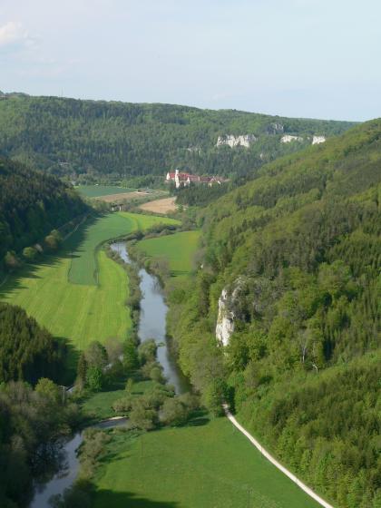 Blick aus großer Höhe auf eine schmale Talaue mit Flussschlinge. Rechts des Gewässers sowie im Hintergrund erheben sich bewaldete Berghänge mit einzelnen hervortretenden Felsen.
