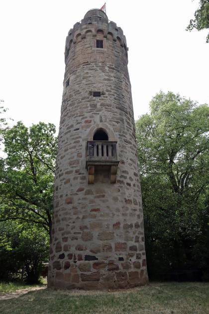 Das Bild zeigt einen hohen gemauerten Rundturm mit Krone und Aufsatz. In der Bildmitte befindet sich eine größere Öffnung im Turm mit vorgesetztem Balkon. Der Turm steht auf einer Lichtung; er überragt die Bäume, die dahinter stehen.