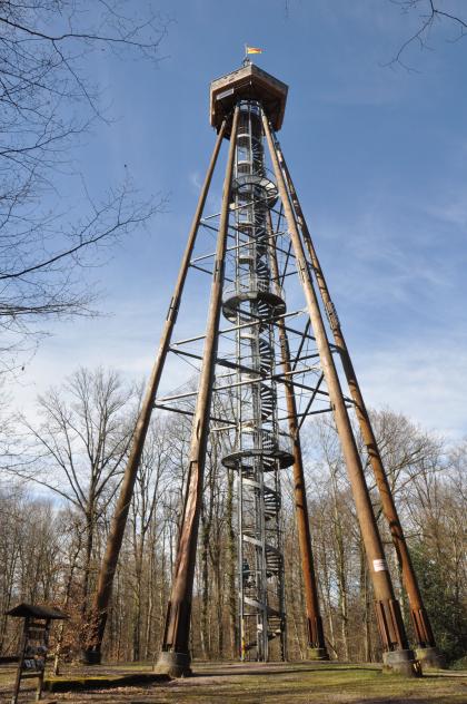 Auf einer Lichtung steht ein von Bäumen umringter Aussichtsturm. Der Turm besteht aus mehreren, oben zusammenlaufenden Holzstämmen. In der Mitte ist eine Wendeltreppe aus Eisen angebracht, die - gesäumt von mehreren Plattformen - hinaufführt.