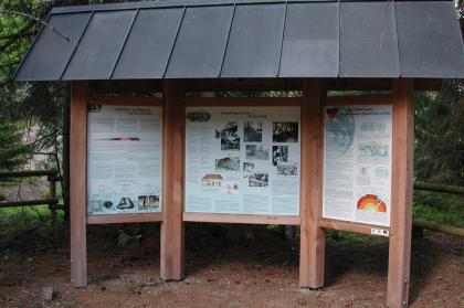 Das Bild zeigt drei durch hölzerne Streben verbundene Schautafeln. Die mittlere Tafel erzählt von der Industriegeschichte im Hotzenwald, die beiden anderen von Gesteinen und Erdgeschichte. Alle Tafeln sind überdacht.