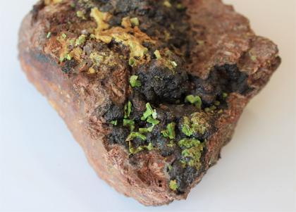 Großaufnahme eines unten dreieckigen Gesteinsstückes. Die rötlich braune Oberfläche weist schwarzgraue Knollen auf, an denen kleine grüne Kristalle sitzen.