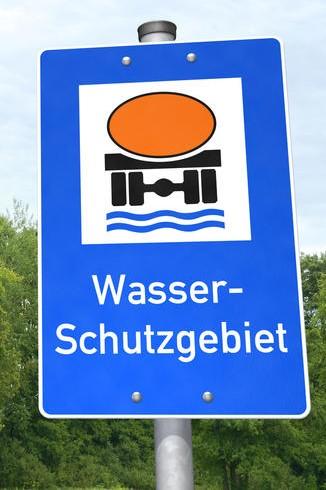 Das Bild zeigt ein blaues Schild mit weißer Schrift, das auf ein Wasserschutzgebiet hinweist. Durch einen stilisierten Gefahrgut-Lkw wird außerdem zur besonderen Vorsicht gemahnt.