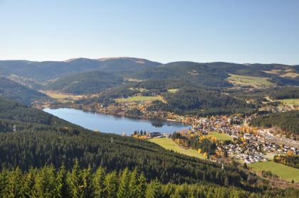 Von erhöhtem Standpunkt aus blickt man auf eine bergige und bewaldete Landschaft mit einem länglichen See in der Bildmitte. Rechts des Sees schließt sich eine größere Siedlung an.