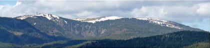 Panoramabild von bewaldeten, nebeneinander liegenden Bergen mit von Schnee bedeckten Gipfeln. Auf dem linken, höchsten Gipfel ist ein Aussichtsturm erkennbar.