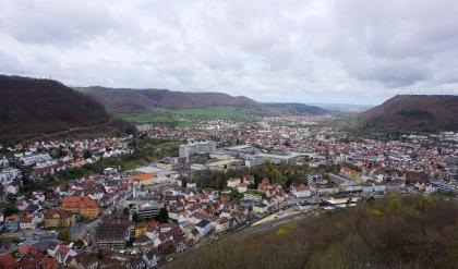 Aus größerer Höhe schaut man auf eine große Stadt, die sich im Talgrund zwischen den Ausläufern bewaldeter, abgeflachter Berge links und rechts hindurchschlängelt.
