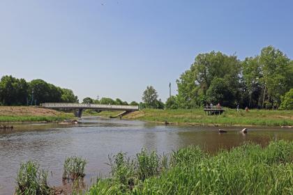 Das Bild zeigt das Zusammentreffen zweier Flußläufe mit flachen bis leicht erhöhten Uferbereichen und Bewaldung im Hintergrund. Der Fluss links wird von einer flachen Brücke überspannt. Am Ufer des Flusses rechts befindet sich eine Aussichtsplattform.