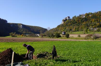 Das Bild zeigt das Aufgraben eines Bodenprofils inmitten einer grünen Wiese. Im Hintergrund sind jeweils zu den Rändern hin ansteigende bewaldete Hänge erkennbar; links mit herausragenden Felsen, rechts mit einer Burg.