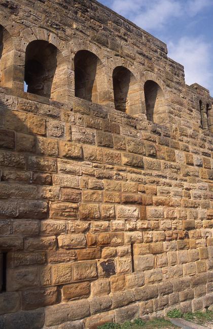 Das Foto zeigt eine hoch aufragende, rötlich graue Steinmauer mit fensterartigen Durchlässen im oberen Bereich.