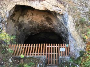 Abwärts gerichteter Blick auf den Eingang einer Höhle. Die Höhle, die sich am Ende eines Felshanges befindet, ist mit einem hölzernen Gitter sowie einer Tür verschlossen.
