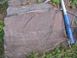 Nahaufnahme eines rötlich grauen Gesteinsbrockens mit hellerem Streifen oben links. Rechts lässt der Stiel eines Hammers die Größe ahnen.