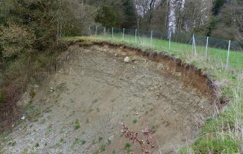 Blick auf einen nach links abfallenden, oben durch einen Zaun gesicherten kiesigen Hang. 