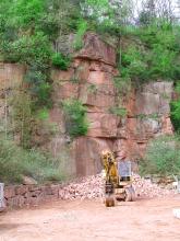 Das Bild zeigt eine stark zugewachsene, rötlich graue Steinbruchwand. Im unteren Bildteil sind Abraumsteine aufgehäuft sowie ein Bagger abgestellt worden.