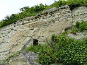 Das Bild zeigt eine hohe, nach rechts ansteigende Felswand mit waagrecht verlaufenden Furchen und Nischen im oberen Drittel. Am Fuß der Felswand ist eine Höhle erkennbar. Rechts unten ist das hellbraune bis graue Gestein bewachsen, ebenso auf der Kuppe.