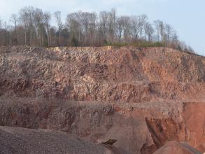 Das Bild zeigt eine mehrstufige Abbauwand von rötlichem Gesteinsmaterial. Auf der Kuppe der Abbauwand sind Bäume zu sehen.