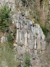 Das Foto zeigt eine auffallende, hohe Felsformation an einem Berghang. Die Formation besteht aus mehreren, durch Klüfte getrennte Felsen, die auf- und nebeneinanderstehen. Teils sind die Felsen auch abgerundet. Der Berghang selbst ist bewaldet.