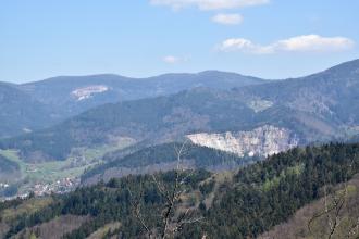 Blick aus großer Höhe auf Reihen bewaldeter Berge. Im Mittelgrund rechts sowie links im Hintergrund sind Steinbrüche erkennbar. Links unten liegt eine Siedlung.