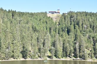 Das Foto zeigt das Ufer eines Sees. Es besteht aus einem steil aufragenden Hang mit dichtem Nadelwald. Auf der Kuppe des Hanges, oberhalb der Bildmitte, ist eine Lücke zwischen den Bäumen. Dort stehen Gebäude. Eines hat einen Aussichtsturm.
