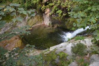 Blick von oben auf ein viereckiges, mit Wasser gefülltes Steinbecken. Der hintere Rand wird von Felswänden gebildet. Rechts fließt Wasser über eine Gefällstufe in das Becken.