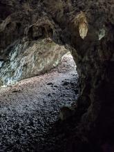 Das Bild zeigt das Innere einer Höhle im Bereich des Ein- beziehungsweise Ausgangs. Der vom linken Bildrand zur Bildmitte verlaufende schmale Gang wird bogenförmig von Gestein überspannt.