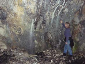 Blick auf eine Höhlenwand mit unterschiedlichen Farben: gelblich (links) bis dunkelgrau (rechts). Eine Besucherin weist rechts am Bildrand auf weißliche, streifenförmige Sinterbildungen in der Höhle hin.