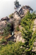 Blick auf steil emporragende, stufig angeordnete rötlich graue Felsen. Der höchste Punkt befindet sich rechts oben. Gleich links darunter steht ein einzelner Baum auf der Felskrone. Links unten ist ein offener Durchgang, ein Felsentor, erkennbar. 