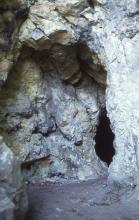 Blick auf ein aus graublauem Gestein gebildetes Felsentor, das den Eingang zu einer sich rechts nach einer Felswand öffnenden Höhle freigibt.