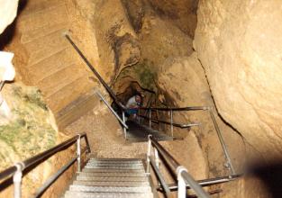 Blick von oben über Metall- und Steintreppen, die steil nach unten in den schmalen Gang einer Höhle führen. Ein Mann mit einer Taschenlampe schaut von dort unten herauf.