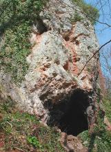 Blick auf eine senkrecht aufragende Felswand. Im hellgrauen, von vertikalen rötlichen Streifen durchzogenen Gestein ist unten rechts eine Höhle oder Nische erkennbar.