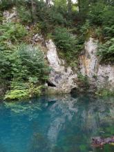 In einer türkisblauen Wasserfläche im Vordergrund spiegeln sich graue, einen Hang bildende Felsen sowie Bäume und Sträucher. Am unteren Ende der Felsen sind kleine Höhlen erkennbar. 