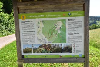 Blick auf eine große farbige Hinweistafel. Die Tafel, die Fotos, eine Karte sowie Informationen zum Castellbergpfad zeigt, steckt in einem Holzrahmen.