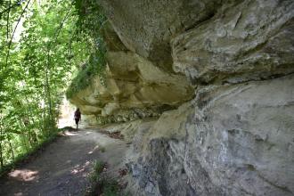 Das Bild zeigt rechts mächtiges, teils wie ein Dach überhängendes gelblich graues Felsgestein. Links, an der Felsformation entlang, verläuft ein schmaler, von Bäumen gesäumter Wanderweg.