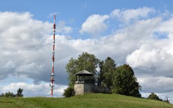 Im Bild ist ein kleiner gemauerter und überdachter Pavillion zu sehen. Der Aussichtsplatz steht auf einer begrünten Bergkuppe und ist von ein paar Bäumen umgeben. Links vom Pavillon steht ein rot-weiß-gestreifter Sendemast.
