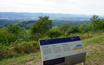 Das Foto zeigt eine mehrfarbige, geknickte Infotafel aus Metall zur Geologie des Hohenstaufen bei Göppingen. Die Tafel steht an einem Aussichtspunkt mit weitem Blick auf die Landschaft.