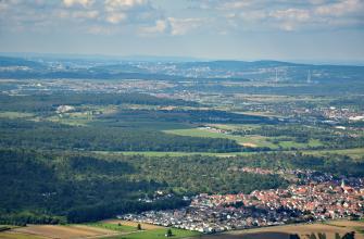 Blick von einem Aussichtspunkt über eine weite, leicht nach links hin ansteigende Landschaft, in der sich Wiesen, Äcker, Waldgebiete und zahlreiche Siedlungen abwechseln. Im Hintergrund sind bewaldete Berge sowie ein eingeschnittenes Tal erkennbar.