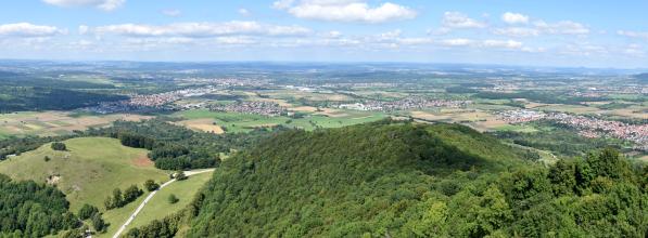 Panoramablick von einem Aussichtspunkt. Über eine bewaldete Bergkuppe im Vordergrund geht der Blick über eine weite flache Landschaft mit Wiesen, Äckern, Waldinseln und Siedlungen.