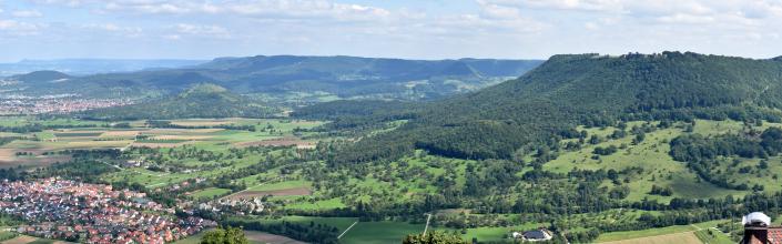 Panoramablick von einem Aussichtspunkt. Vor abgeflachten, bewaldeten Bergen rechts breiten sich Wiesen, Äcker und Siedlungen aus. Links im Hintergrund sind kleinere Vulkanberge erkennbar; dahinter verlaufen bewaldete Höhenzüge.