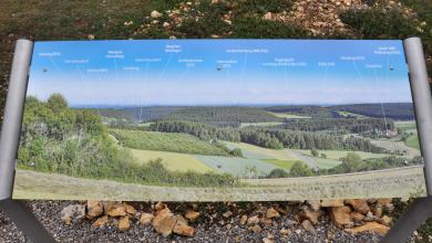 Blick auf eine farbige, an einem Metallrahmen befestigte Fototafel. Die Tafel zeigt eine hügelige Waldlandschaft. Hinzugefügte Namen von Bergen, Ortschaften oder Sehenswürdigkeiten dienen als Orientierungshilfe.