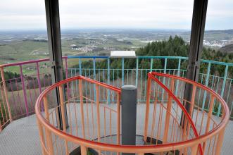 Das Foto zeigt eine Aussichtsplattform mit mehrfarbiger Brüstung aus metallenen Streben. Im Vordergrund der runde Handlauf des Treppenaufgangs. Im Hintergrund eine weite Landschaft mit Wäldern und Weinbergen.