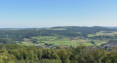 Blick aus größerer Höhe über Baumwipfel auf eine langgestreckte, flache Erhebung im Bildmittelgrund. Die Kuppe der Erhebung ist grün, die Ränder und Hänge sind bewaldet. Daran anschließend folgen von Straßen durchzogene Wiesen sowie einzelne Äcker.