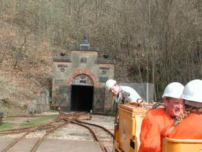 Blick auf den geöffneten, an einem Waldhang gelegenen Eingang eines Bergwerks. Im Vordergrund liegen Schienen der Grubenbahn.