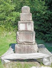 Blick auf ein steinernes Denkmal zu Ehren des Dichters Grimmelshausen. Das Denkmal besteht aus mehreren Steinblöcken unterschiedlicher Größe, die - aufeinandergesetzt - eine Pyramide in Treppenform bilden. Um die Steine herum sind Holzbänke angebracht.