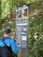 Am Fuße eines steilen, überwucherten Waldhanges steht eine metallene Informationstafel, die das Thema „Die Römer“ hat. Ein Besucher mit Mütze und Jacke sieht sich gerade die Tafel an.