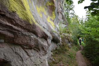 Seitlicher Blick auf eine hohe, rötlich graue Felswand. Das Gestein ist verwittert und zeigt im oberen Bildbereich gelbe Verfärbungen. Rechts führt ein Wanderpfad an der Felswand entlang.